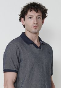 Námořnická modrá polo košile s texturovaným síťováním, kontrastním límcem a krátkými rukávy. Na hrudi se nachází logo.