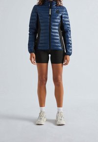 Laivastonsininen toppatakki, jossa on korkea kaulus, vetoketju edessä ja mustat sivupaneelit. Päällä mustat shortsit ja vaaleat lenkkarit.