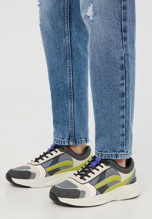 Osoba w niebieskich podartych dżinsach i szaro-czarno-biało-zielonych siateczkowych trampkach stoi na białej powierzchni.