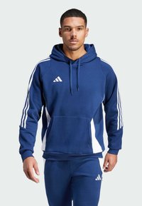 Tamsiai mėlynas džemperis su priekinėmis kišenėmis, baltais šoniniais džemperio kraštais ir trimis juostelėmis ant rankovių. Yra su raišteliu reguliuojama kapuce ir Adidas logotipu.