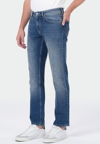 Homme portant un jean droit bleu délavé et des baskets blanches, se tenant avec une main dans sa poche contre un fond uni.