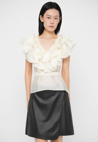 Sieviete ar taisniem melniem matiem, ģērbusies caurspīdīgā baltā blūzē ar volāniem un melnā ādas svārku līdz ceļgaliem, skatās uz priekšu uz baltas fona.