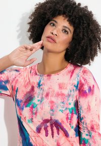 Bunt gemustertes Kurzarmoberteil mit einem pinken und blauen abstrakten Druck, das auf der Vorderseite glitzernde Details in einem kreisförmigen Muster aufweist. Glatte Stoffstruktur.