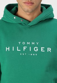 Hanorac de bumbac verde cu buzunar frontal, având un logo alb proeminent "TOMMY HILFIGER EST. 1985" imprimat pe piept.