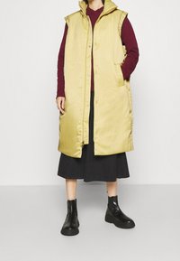 Gul vadderad väst med huva, tryckknappar och sidofickor, bärs över en vinröd hoodie och en svart kjol, i kombination med svarta ankelskor.