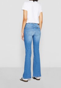 Blue denim flared jeans with a medium wash, mid-rise fit, and two back pockets. Paired with a plain white t-shirt and striped shoes.