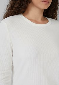 Weißes, geripptes Langarmshirt mit Rundhalsausschnitt. Der Stoff zeigt eine feine Textur mit subtilen vertikalen Linien.