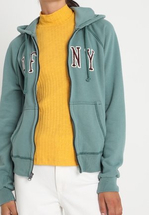 Personne portant un sweat à capuche vert zippé avec des écussons lettre et un pull côtelé jaune, porté avec un pantalon blanc, sur un fond uni.