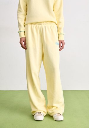 Osoba nosząca luźne, jasnożółte spodnie dresowe i pasującą bluzę, stoi na zielonej podłodze, widoczne białe trampki.