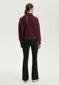 Pulover tricotat de culoare burgundă, cu guler și model texturat, purtat cu pantaloni evazați negri și încălțăminte de nuanță deschisă.