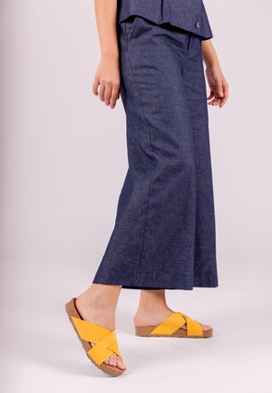 Pantalon large bleu foncé en tissu texturé, associé à des sandales à lanières croisées jaunes avec une semelle en liège et un talon plat.
