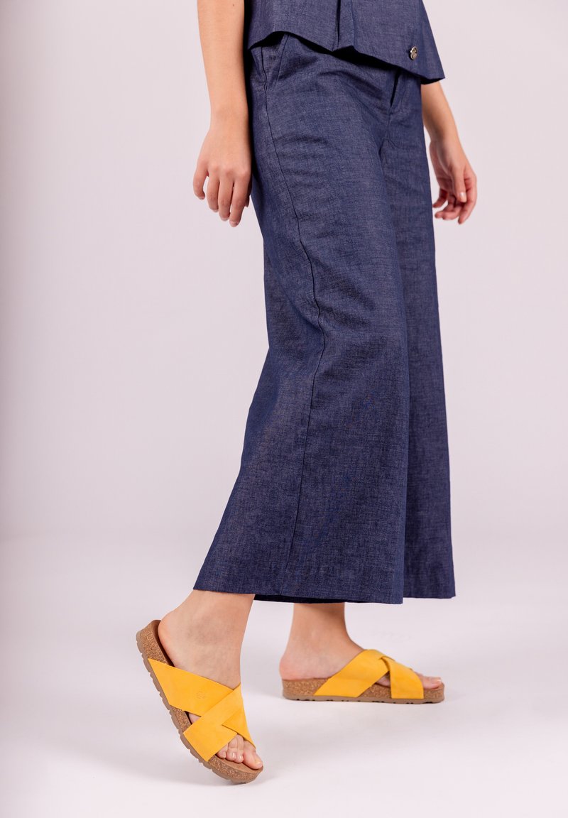 Pantalones de pierna ancha de color azul oscuro, confeccionados con una tela texturizada, combinados con sandalias amarillas de tiras cruzadas, con suela de corcho y tacón plano.