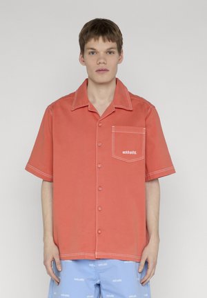 Mladić nosi široku koraljnu košulju na kopčanje s bijelim prošivima i džepom na prsima, uparenu sa svijetloplavim kratkim hlačama s bijelim tekstualnim uzorkom.