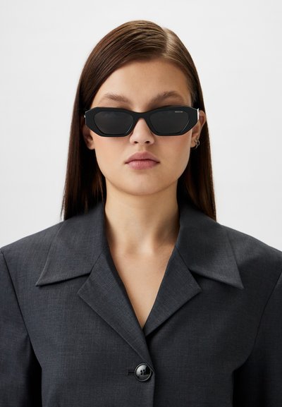 Black sunglasses with an angular shape and dark lenses. Textured, matte frames with subtle branding. Worn with a dark gray blazer.