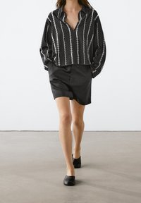 Black patterned shirt with loose fit, featuring white vertical stripes, paired with a black skirt. Model wearing simple black flats.