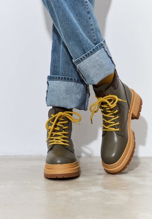Bottes en cuir vertes à la cheville avec des lacets jaunes, semelle en caoutchouc et bout rond. Portées avec un jean bleu retroussé et des accents marron sur le talon.