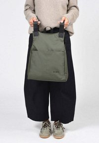 Bolsa tote de nylon verde con dos correas negras, bolsillo con cremallera en el frente y logo de KCB. Diseño minimalista, forma cuadrada y textura mate.