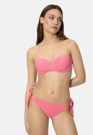 Mujer con bikini rosa sin tirantes y bragas con lazos laterales, de pie con una mano detrás de la espalda, mirando hacia un lado.