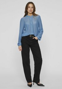 Blouse bleu clair à manches longues avec des détails en dentelle, fermeture à boutons et col rond, associée à un pantalon noir droit et des escarpins noirs.