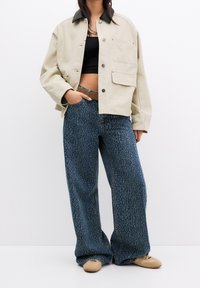 Beige canvas jas met een donkere kraag, gecombineerd met wijde blauwe jeans met een luipaardprint patroon. Tan schoenen maken de look compleet.