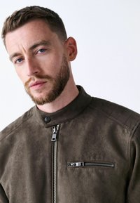 Chaqueta de ante marrón con un alto cuello, cremallera frontal y un bolsillo en el pecho con cremallera. Presenta una textura suave y un sutil brillo.
