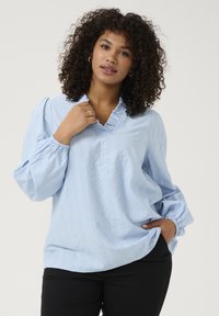 Blouse bleu clair à manches longues avec un col en V et un col froncé, présentant des rayures texturées et des manches regroupées. Associée à un pantalon noir.