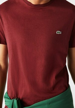 Homme portant un t-shirt bordeaux uni à manches courtes avec un petit logo crocodile vert sur la poitrine gauche et un tissu vert noué autour de la taille.