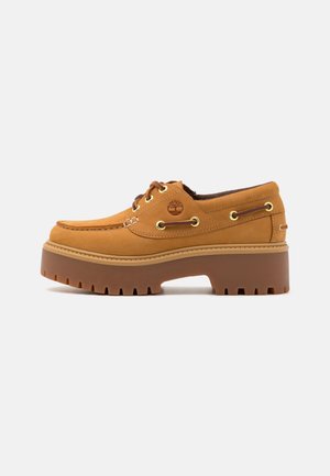 Buty typu boat w kolorze jasnobrązowym, z grubą podeszwą, wiązane z przodu, z metalowymi oczkami, przeszyciem typu moc toe i logo drzewa Timberland po bokach.