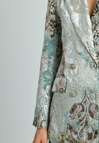 Blazer doppiopetto in tessuto testurizzato teal e argento, con un intricato motivo floreale e due tasche frontali con bottoni.