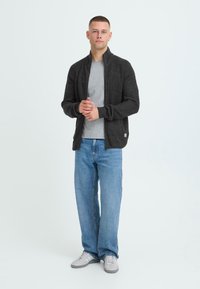 Mørkegrå ribbestrikket cardigan med høy krage, brukt over en lys grå t-skjorte, sammen med lyseblå jeans og grå joggesko.