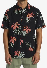 Chemise noire à manches courtes présentant un motif floral tropical en rouge et vert, avec une patte de boutonnage et une poche poitrine.