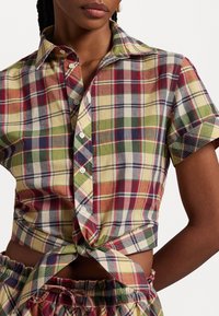 Short-sleeved, collared plaid shirt in yellow, red, and green tones, featuring buttons and a knotted front design.