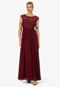 Lungo vestito da sera bordeaux con corpetto in pizzo, maniche a cappuccio, vita arricciata e gonna in chiffon fluida. Semplici tacchi con cinturino alla caviglia completano il look.
