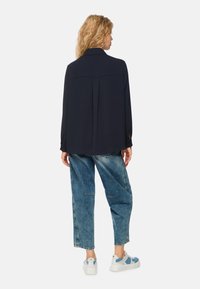 Navy long-sleeve shirt with a collar, featuring a back yoke seam and side slits, paired with light blue denim pants and white sneakers.