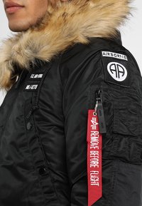 Black bomber jacket featuring a textured fur hood, utility pockets, and a red "REMOVE BEFORE FLIGHT" tag with white text.