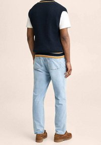 Mann in hellblauen Jeans, braunen Schuhen, einem weißen kurzärmeligen Hemd und einem dunkelblauen Strickwesten mit gelben und weißen Streifen, steht mit dem Rücken zur Kamera.