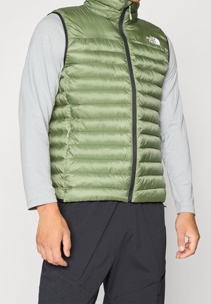 Homme portant un gilet matelassé vert sans manches par-dessus une chemise gris clair à manches longues et un pantalon noir, debout devant un fond uni.