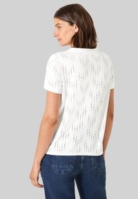 T-shirt bianco a maniche corte con un motivo testurizzato di piccoli ritagli. Collo rotondo e vestibilità ampia. Abbinato a jeans blu scuro.