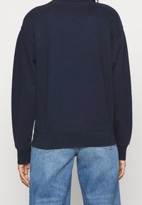 Námořnická modrá mikina s volným střihem, žebrovanými manžetami a lemem, s hladkou texturou. V kombinaci se světle modrými džíny.