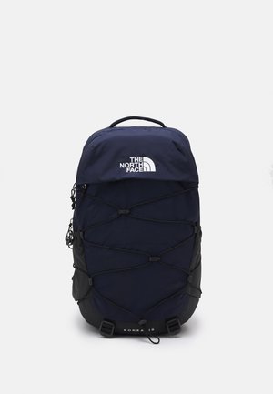 Mochila de color azul marino con detalles en negro, que cuenta con un sistema de cordón elástico, un asa superior y un logotipo frontal. Fabricada con tejido resistente, diseño estructurado.