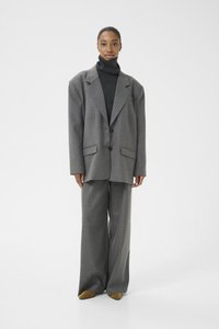 Veste de costume grise surdimensionnée avec de larges revers, associée à un pantalon gris à jambes larges et à un pull col roulé noir, le tout complété par des chaussures jaunes.