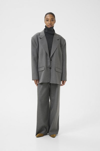 Veste de costume grise surdimensionnée avec de larges revers, associée à un pantalon gris à jambes larges et à un pull col roulé noir, le tout complété par des chaussures jaunes.