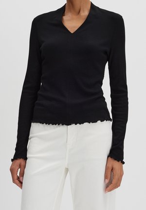 Kobieta ubrana w dopasowaną czarną bluzkę z długim rękawem, dekoltem w serek i falbankami przy krawędziach, w zestawieniu z białymi, wysokim stanem spodniami.