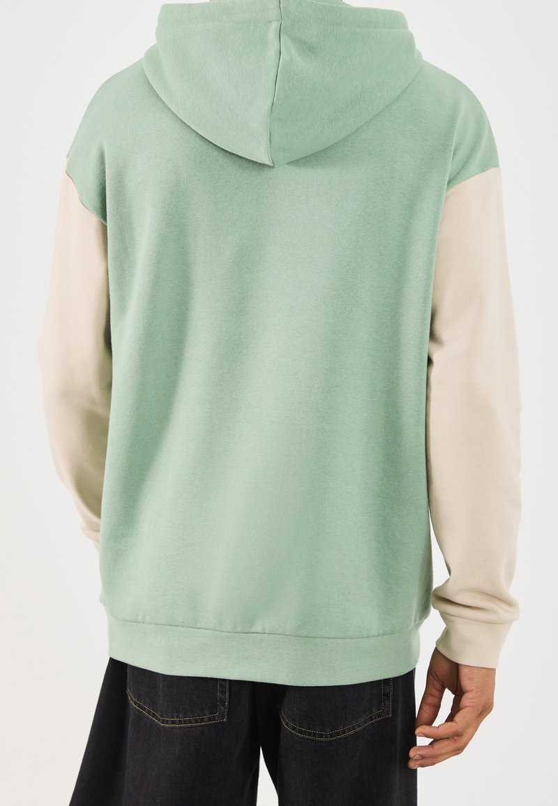 Personne portant un sweat à capuche avec un corps vert sauge et des manches couleur crème, debout dos à la caméra.