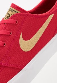 Czerwony płócienny tenisówka z wyszywanym na złoto logo Nike w kształcie łuku, posiadająca białą gumową podeszwę i kontrastowe różowe sznurówki.