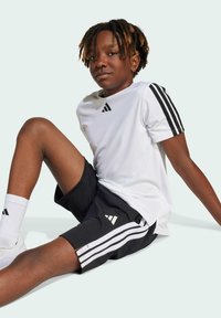 Camiseta deportiva blanca con rayas negras en las mangas, combinada con unos pantalones cortos negros que tienen rayas blancas. Modelo sentado, usando calcetines blancos y zapatillas.
