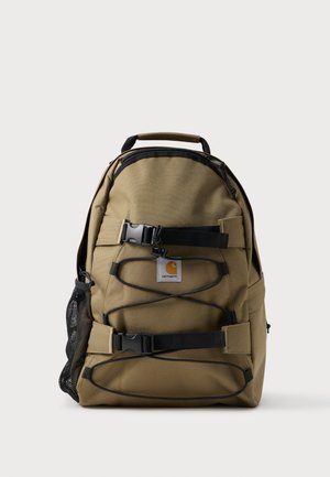 Brun Carhartt rygsæk med sorte justerbare stropper, to spænderlukninger, elastiske snore foran og en sidelomme i mesh.