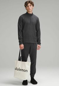 Donkergrijze heren-pullover met kwartrits en een gestructureerd oppervlak, bijpassende joggers en een crèmekleurige tas met "lululemon" in het zwart gedrukt.