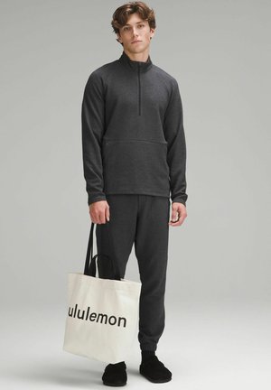 Sudadera de hombre gris oscuro con cremallera hasta el cuarto, superficie texturizada, pantalones jogger a juego y bolsa tote crema con la palabra "lululemon" impresa en negro.