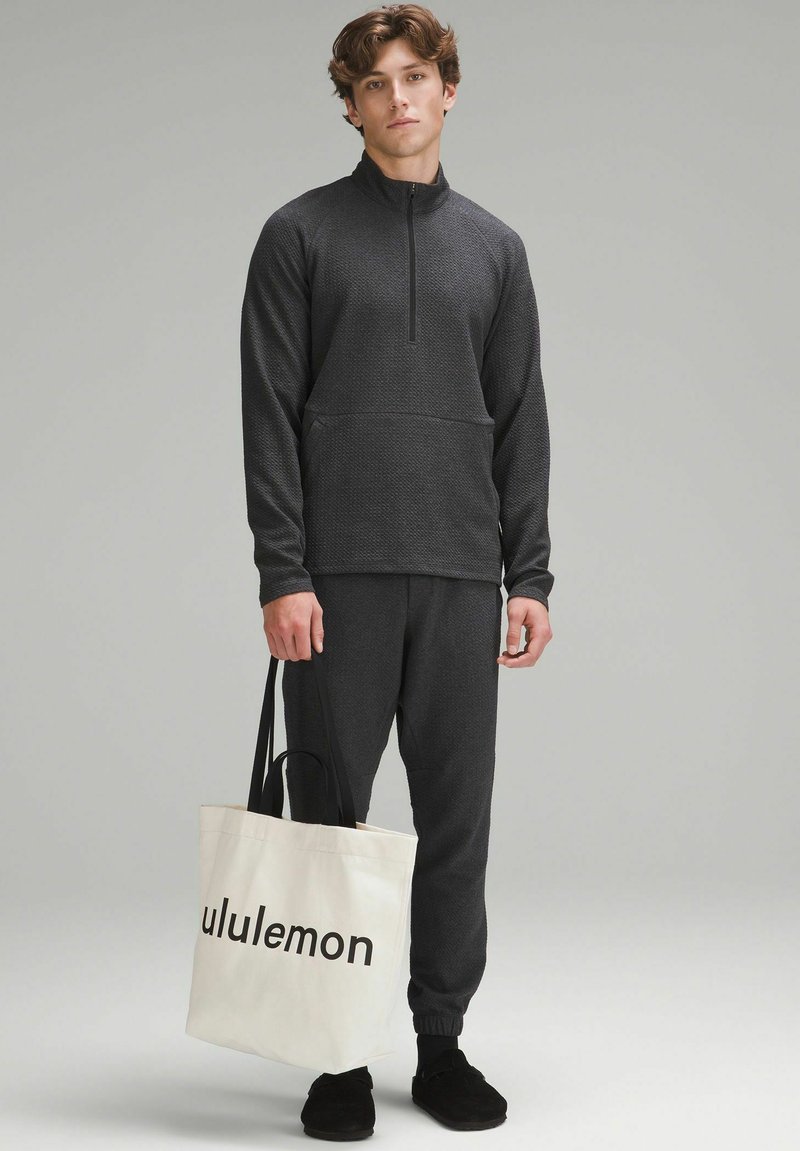 Donkergrijze heren-pullover met kwartrits en een gestructureerd oppervlak, bijpassende joggers en een crèmekleurige tas met "lululemon" in het zwart gedrukt.